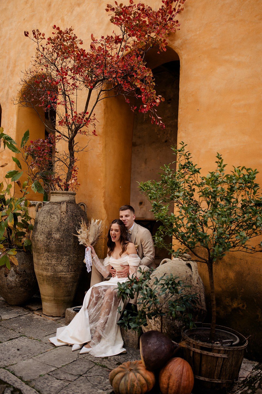 Couple in wedding attire seated against rustic ochre wall with autumn foliage and large terracotta urns.