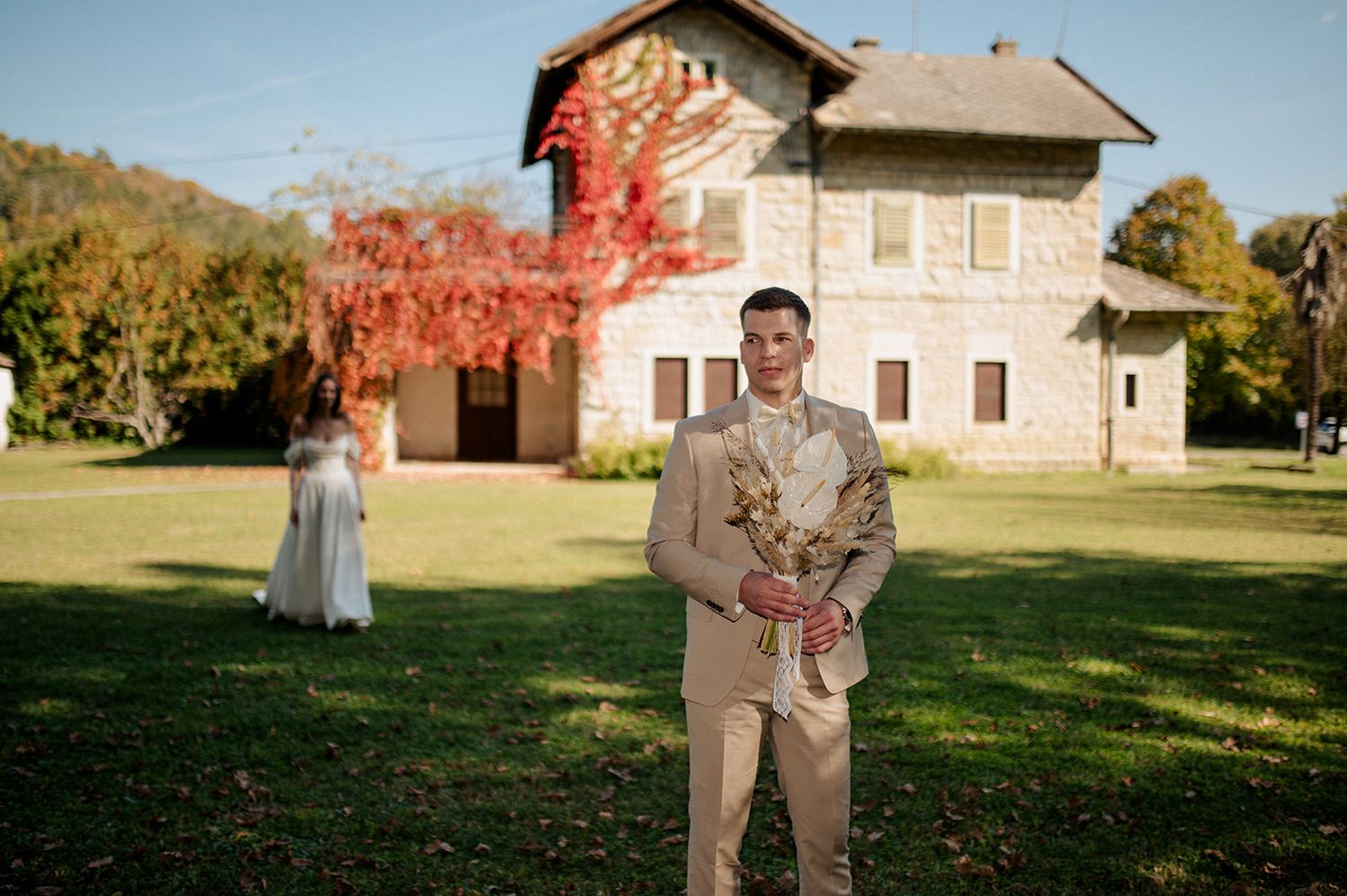 Groom in tan suit holding dried floral bouquet stands on green lawn before stone house with red ivy foliage.