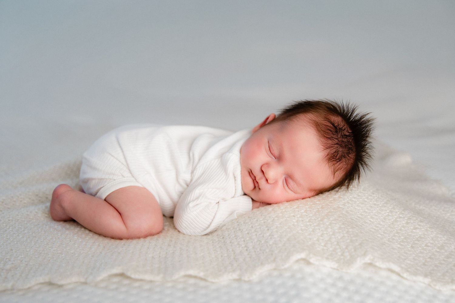 A sleeping baby in white clothing lies peacefully on a cream-colored blanket.