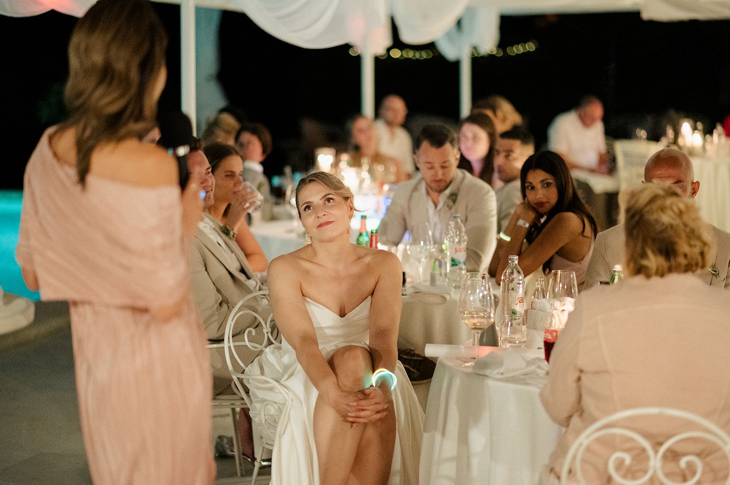 Bride sits at candlelit outdoor wedding reception while guests celebrate at decorated tables.