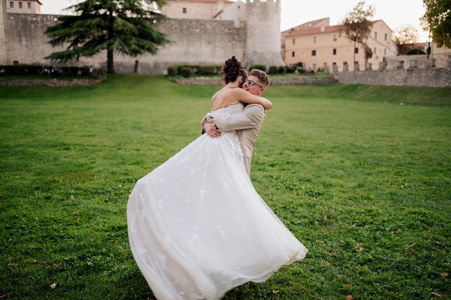 Groom lifting bride in white gown outdoors near historic castle walls.