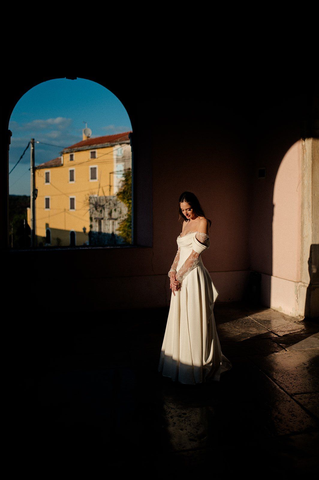 Bride in white off-shoulder gown stands in sunlit archway with yellow building backdrop.