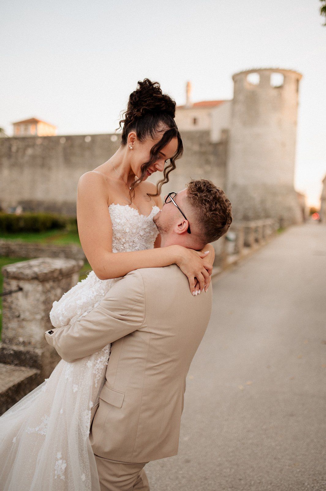 Groom in beige suit lifts bride in white lace wedding dress near ancient stone walls at sunset.