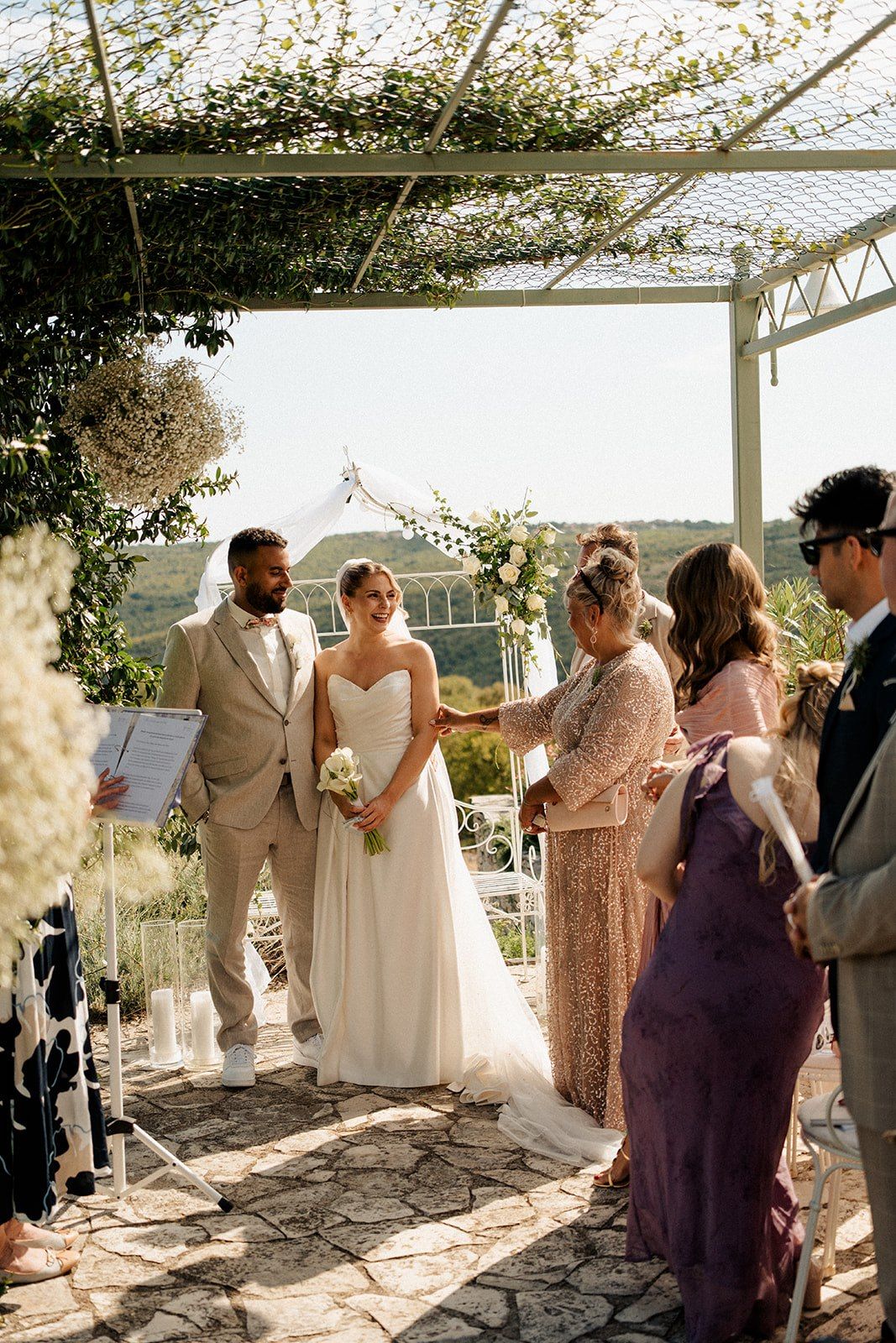 Bride and groom exchanging vows under floral pergola at outdoor wedding ceremony with guests and officiant.