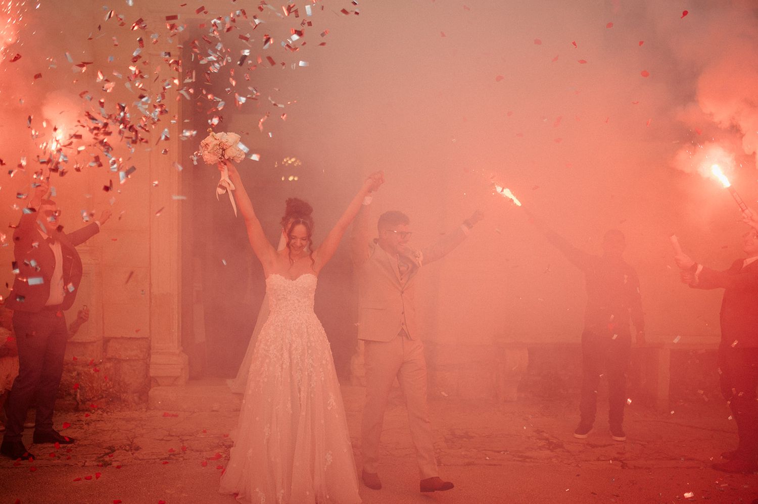 Bride and groom celebrate with red smoke flares and confetti during dramatic outdoor wedding exit at night.