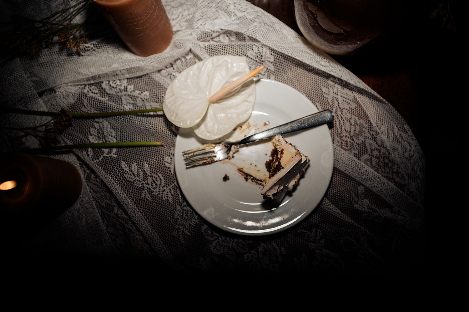 A dimly lit plate with a slice of layered cake, fork, knife, and sliced pear on a lace tablecloth.