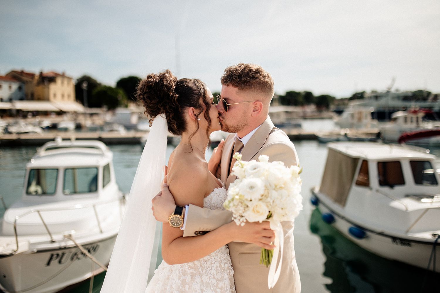 A newlywed couple kissing at a sunny harbor, bride holding white bouquet with boats behind them.