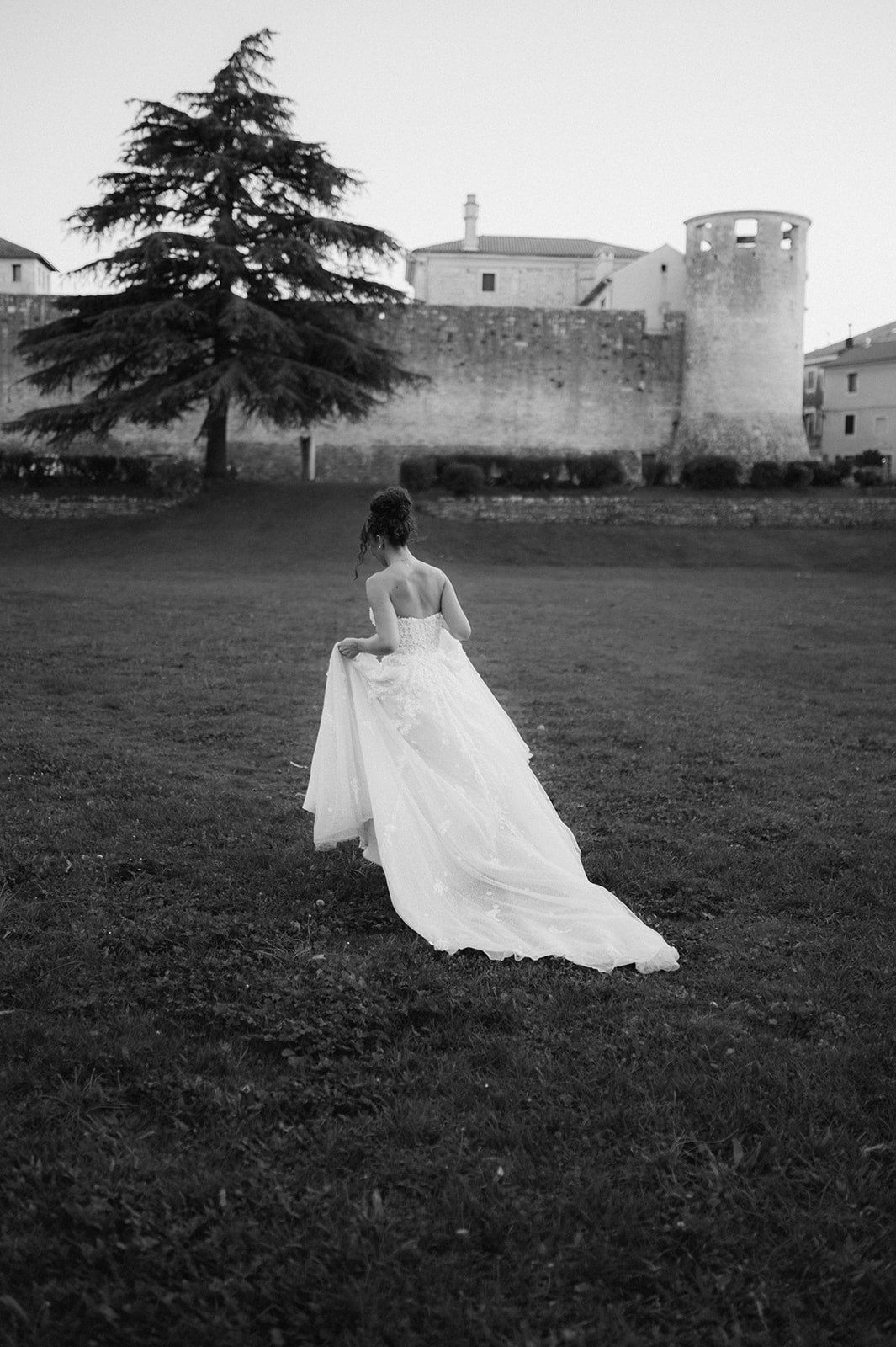 Bride in elegant backless wedding gown with long train walks toward historic stone castle in black and white photo.