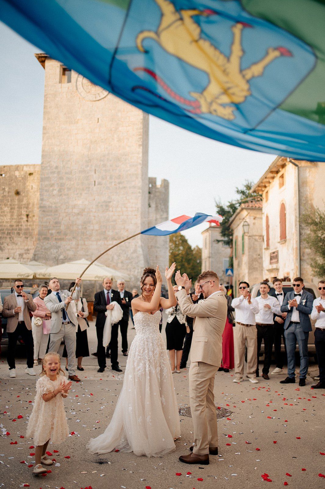 Bride and groom celebrate wedding exit with confetti and Croatian flags outside historic Dubrovnik stone walls.