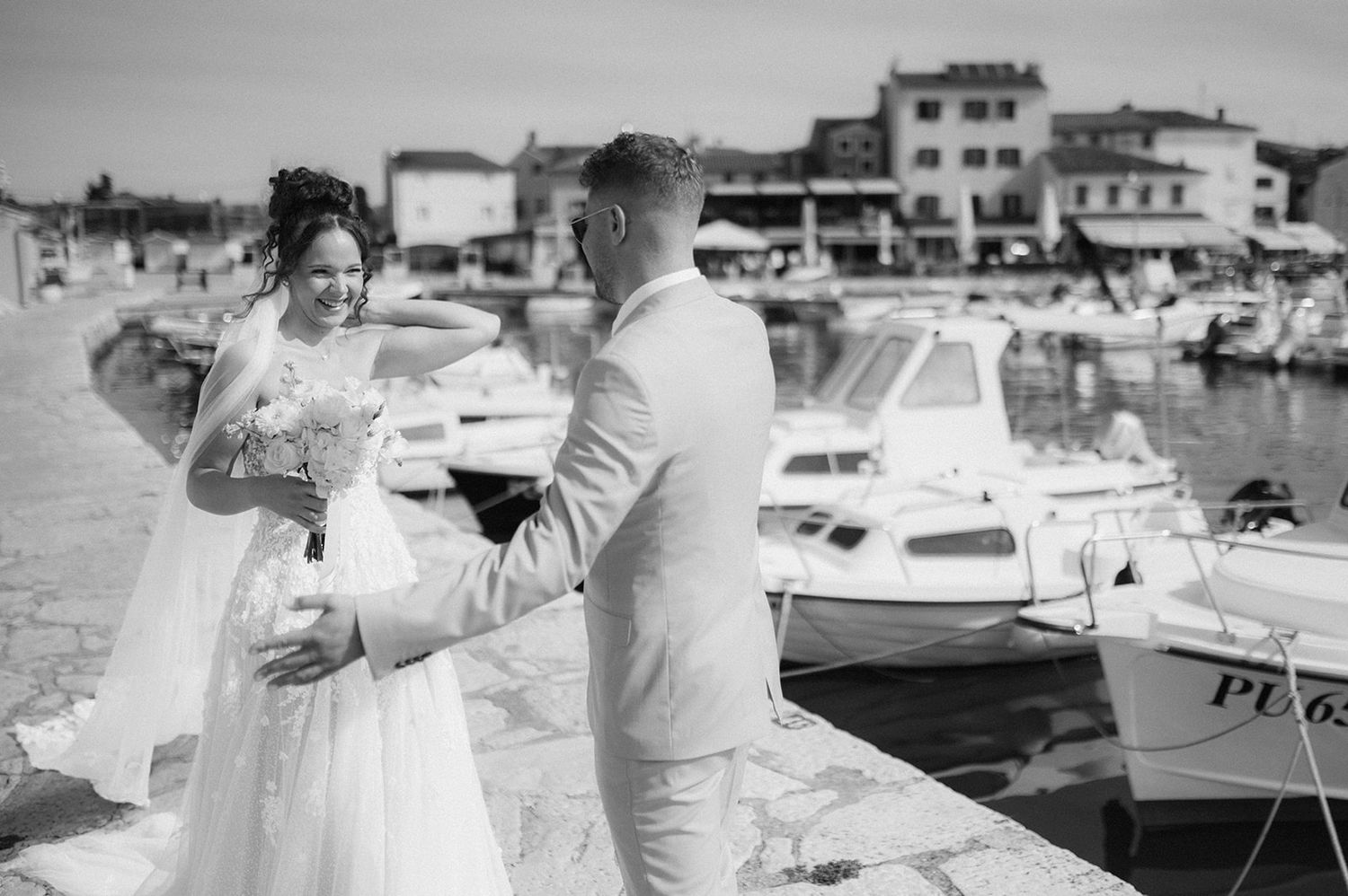 Black and white photo of a bride and groom sharing a tender moment on a marina dock with boats and buildings behind them.