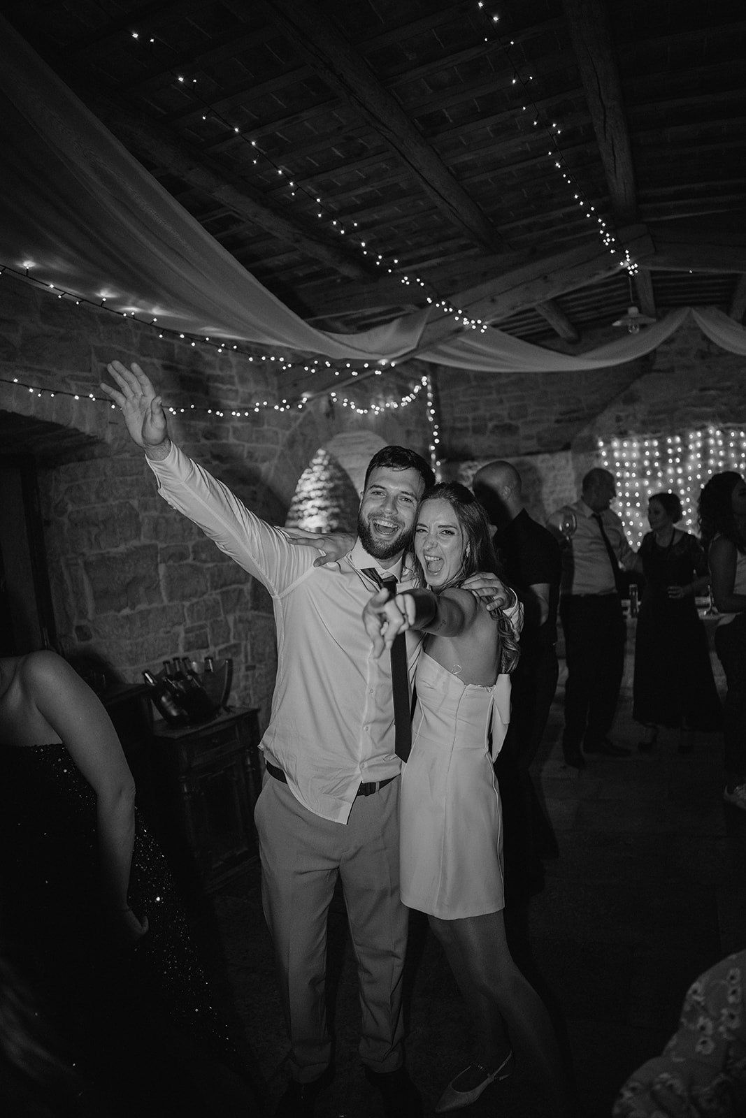 Black and white photo of a joyful couple dancing at a wedding reception with fairy lights and draped fabric overhead.