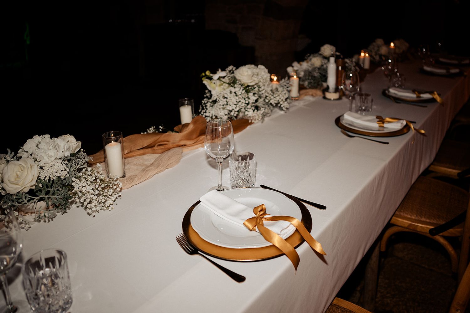Elegant wedding reception table with white floral centerpieces, gold charger plates, candles, and white linen tablecloth.