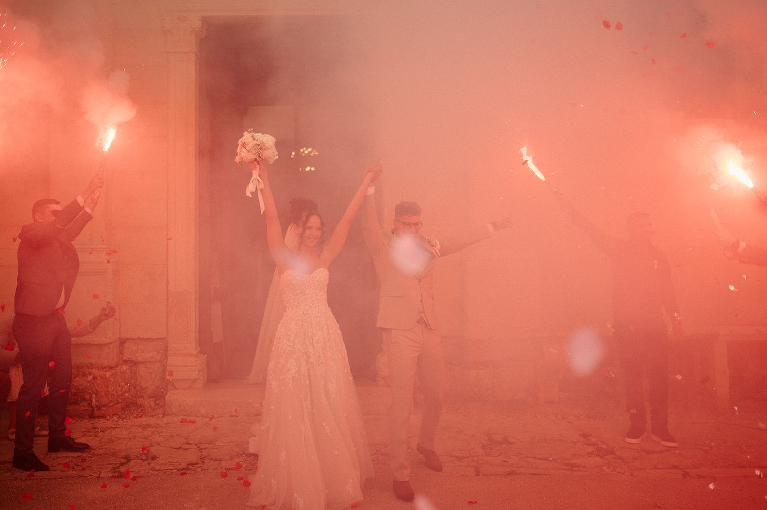 Bride and groom exit ceremony surrounded by red smoke flares held by guests at an outdoor wedding celebration.
