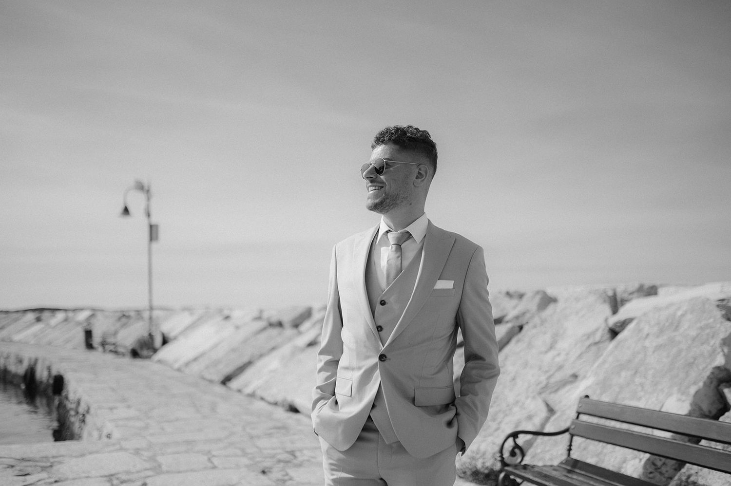 Stylish groom in a light gray suit and sunglasses poses outdoors near a bench in a black and white photo.
