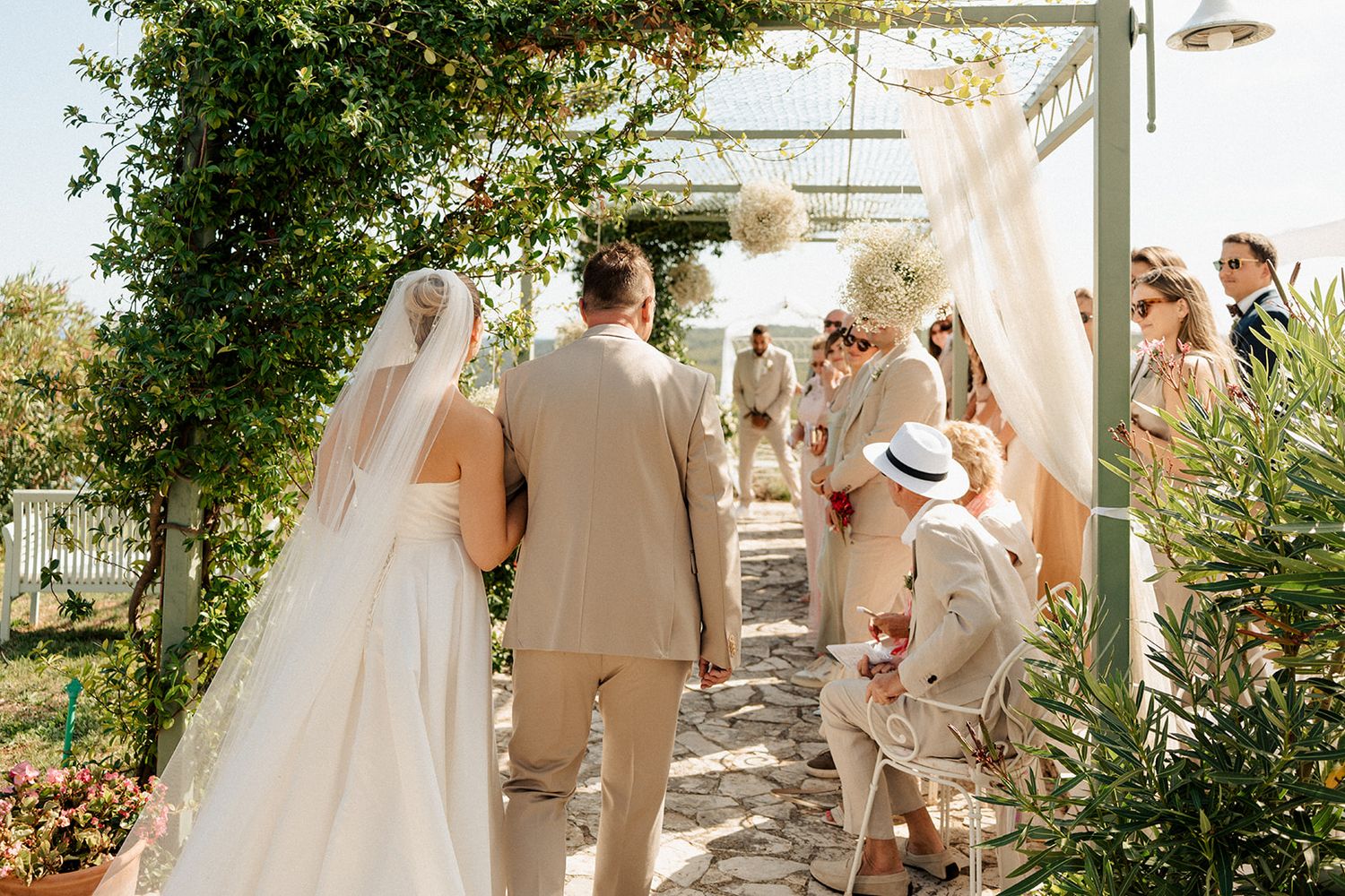Bride and groom walk down outdoor aisle flanked by wedding guests at a sunlit garden ceremony.