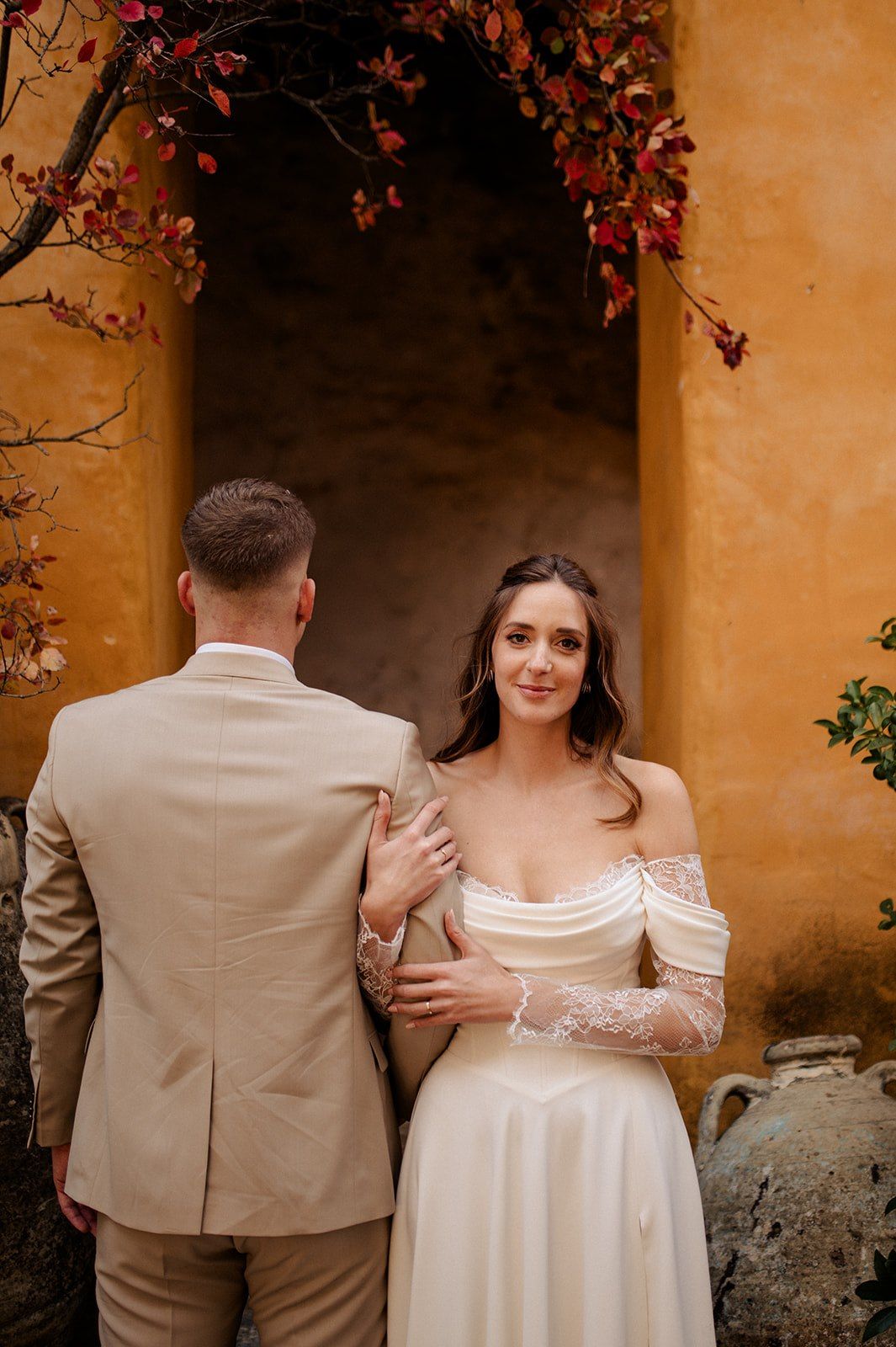 Bride in off-shoulder lace wedding gown with groom in tan suit posing by golden wall with autumn foliage arch.