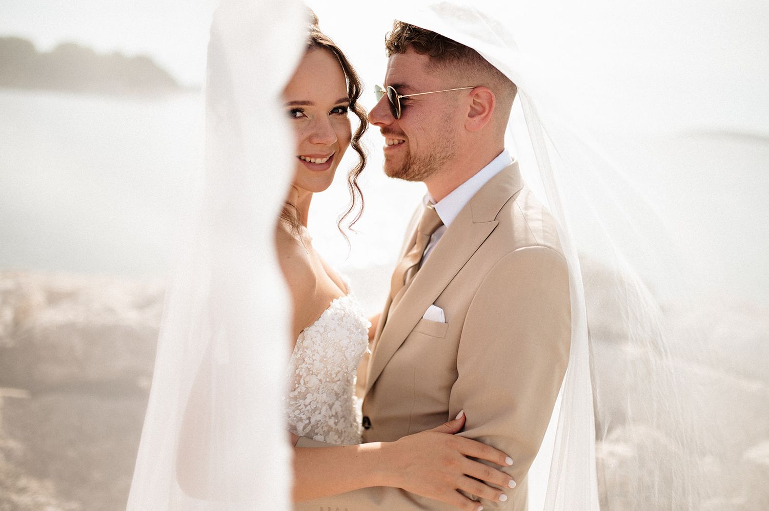 Bride and groom sharing a romantic moment outdoors, wrapped in a white veil, with a beachside backdrop.
