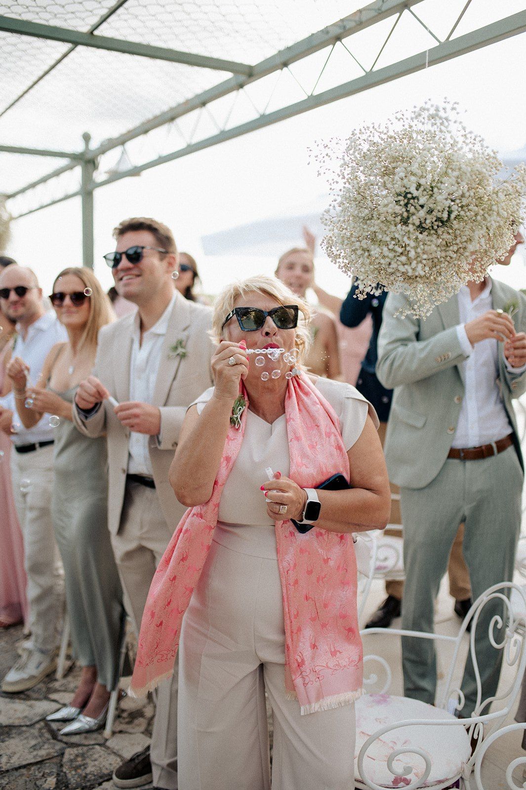 Wedding guests in elegant attire photograph ceremony outdoors, woman in pink scarf holds vintage camera.