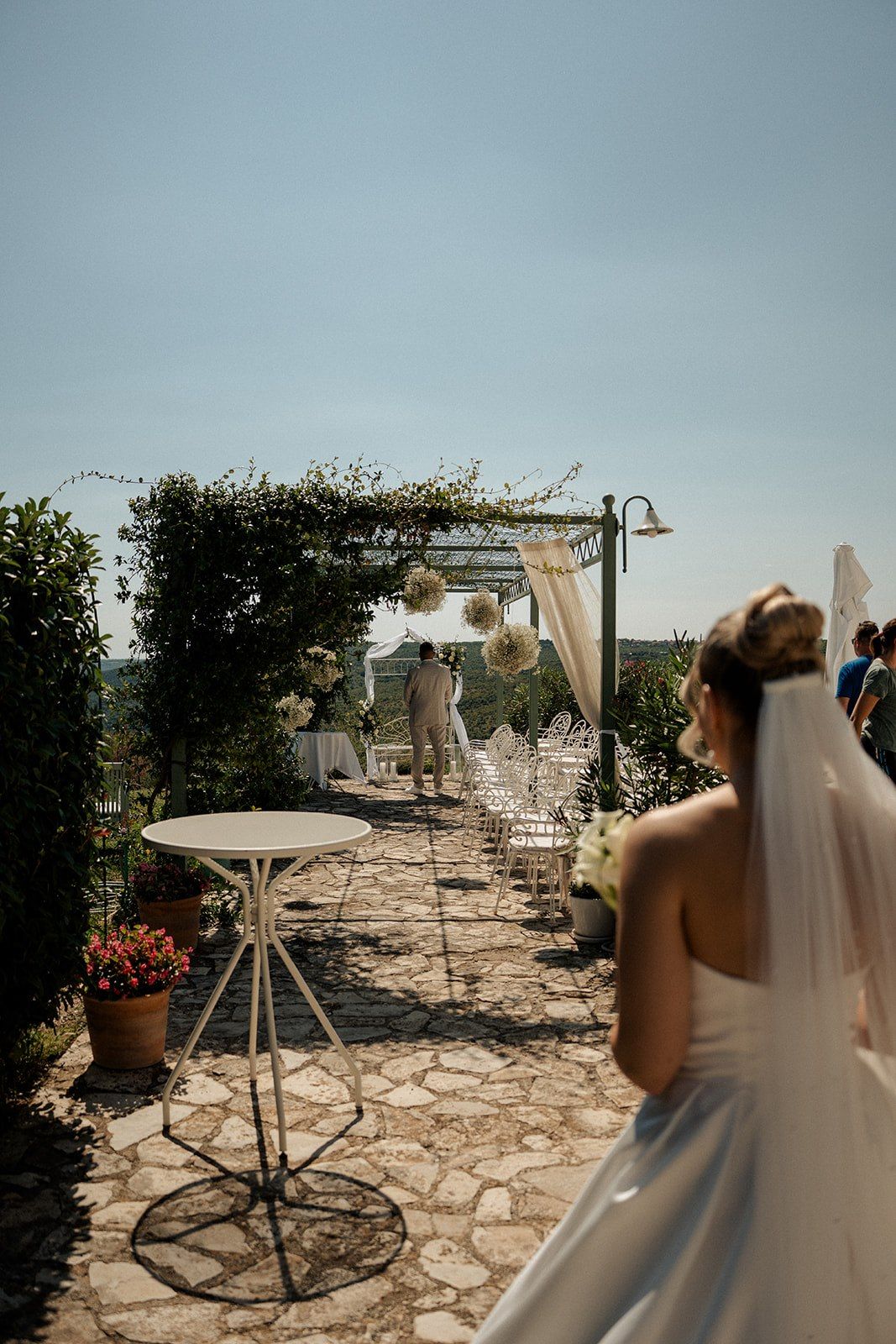 Bride in white gown and veil walks toward outdoor wedding ceremony arch decorated with flowers and draping.