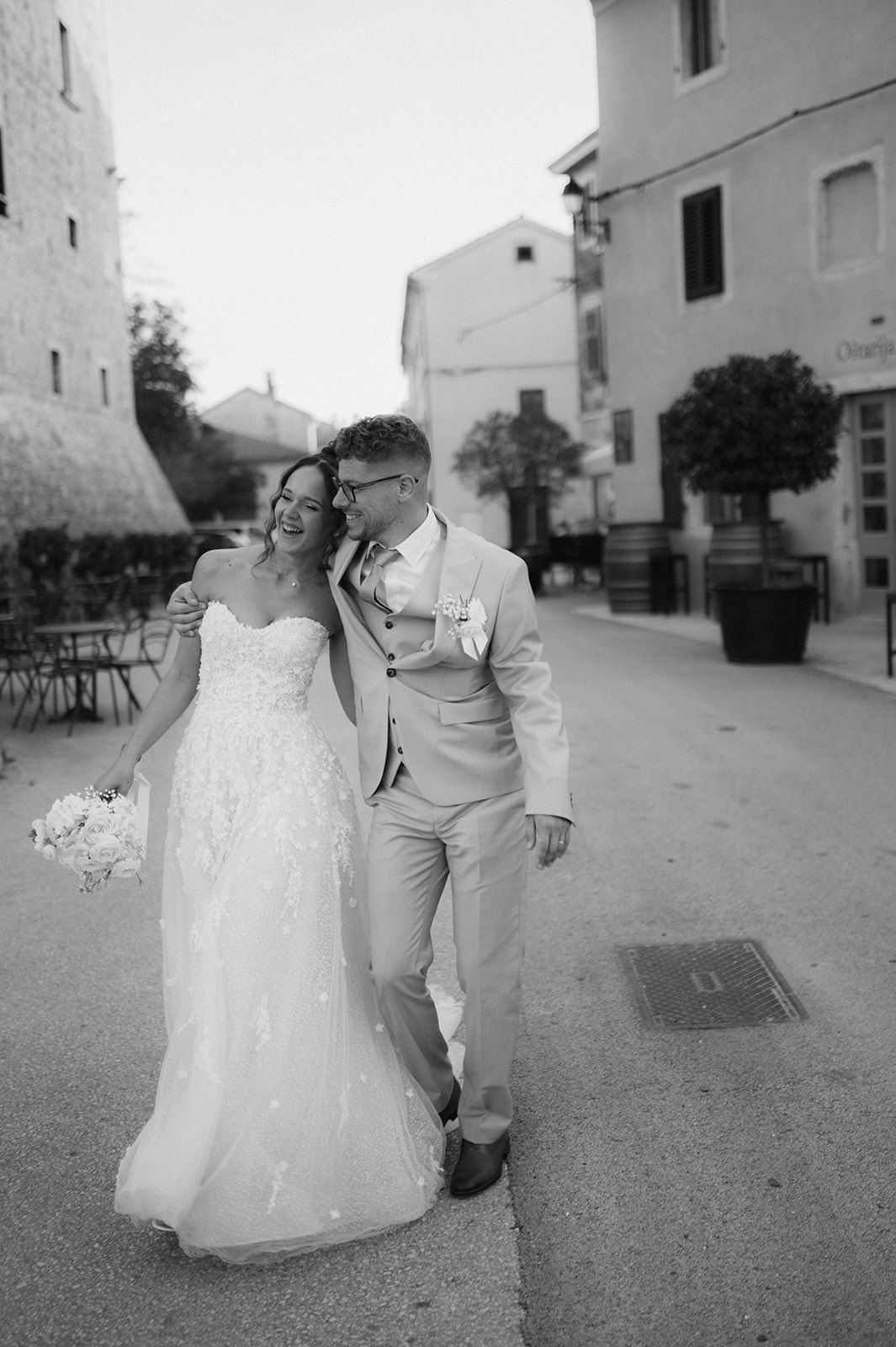 Black and white photo of a smiling bride and groom walking together on a cobblestone street on their wedding day.