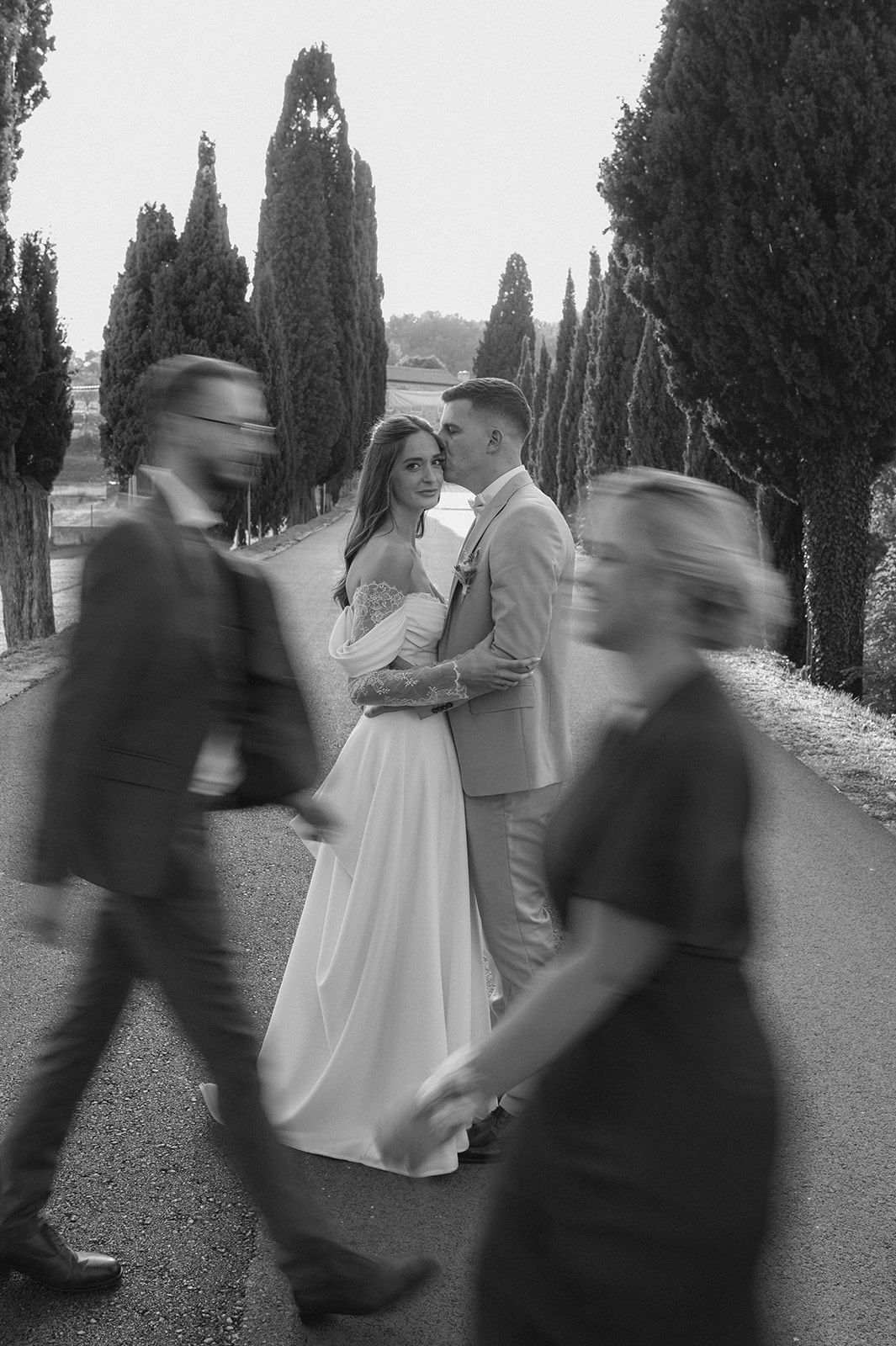 Black and white wedding photo of bride and groom embracing on tree-lined path with blurred guests walking past.