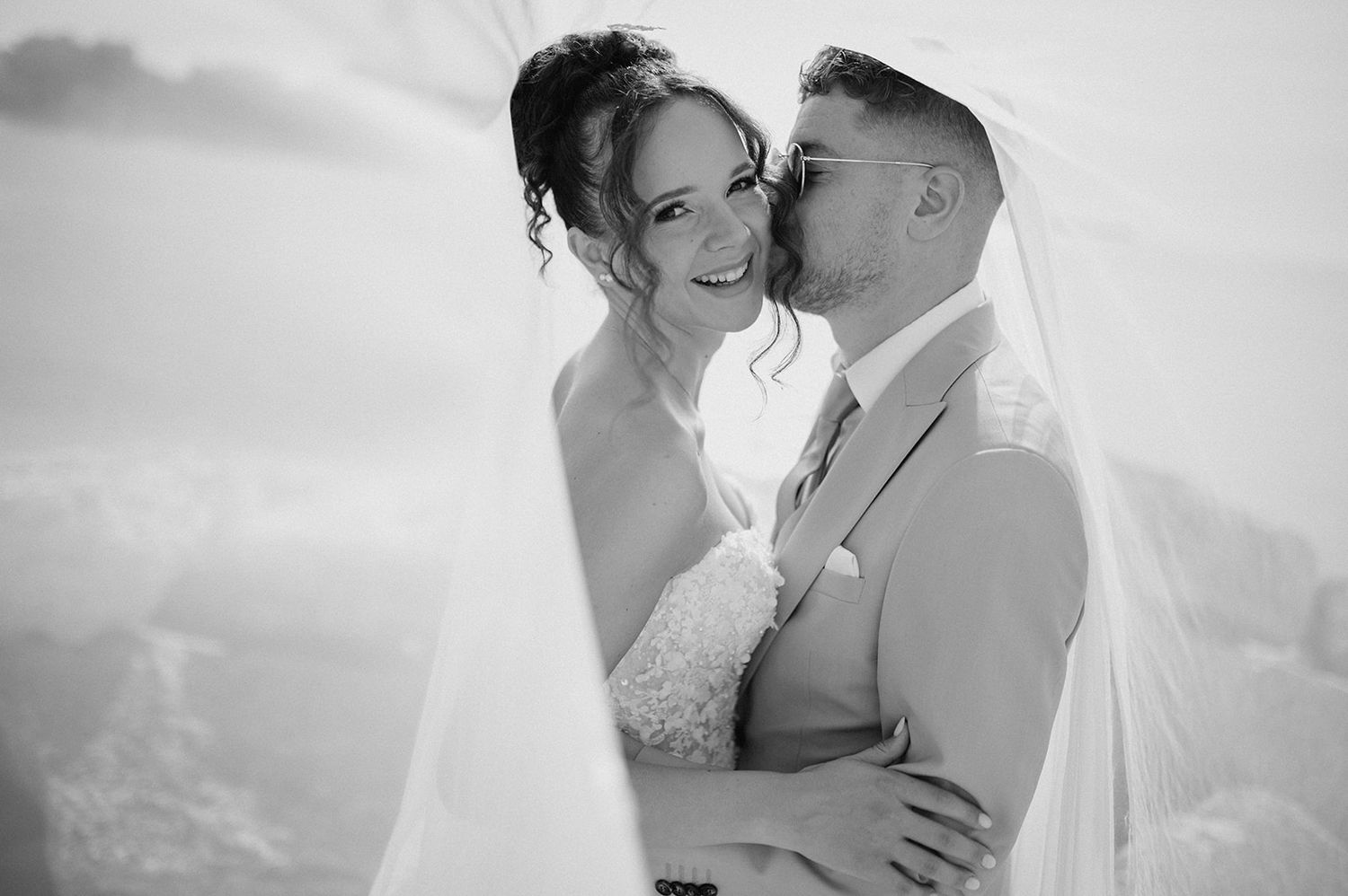 Black and white wedding photo of bride and groom embracing under veil, groom kissing bride's cheek outdoors.