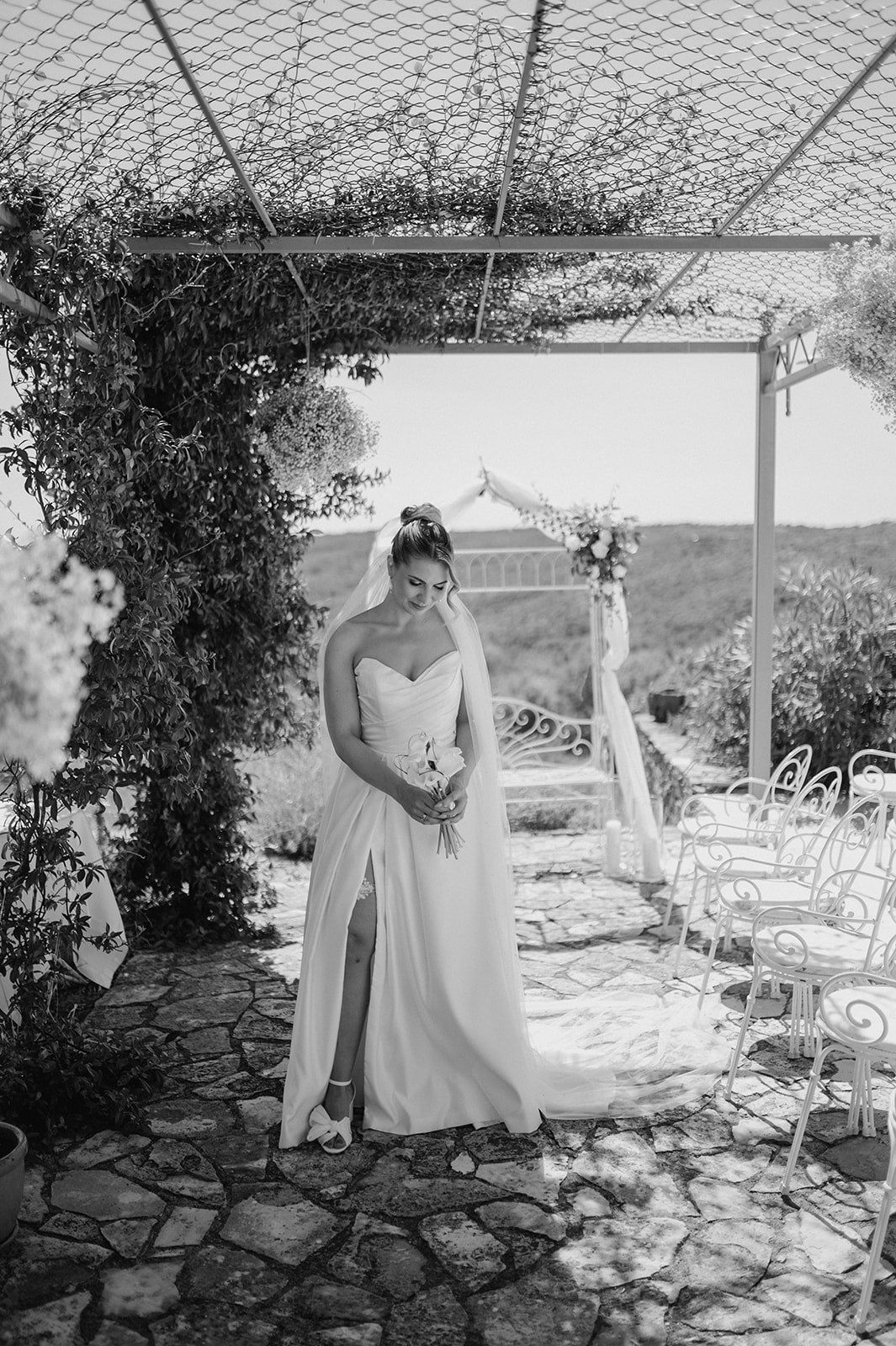Bride in strapless wedding gown with slit and veil stands under floral arch at outdoor ceremony venue.