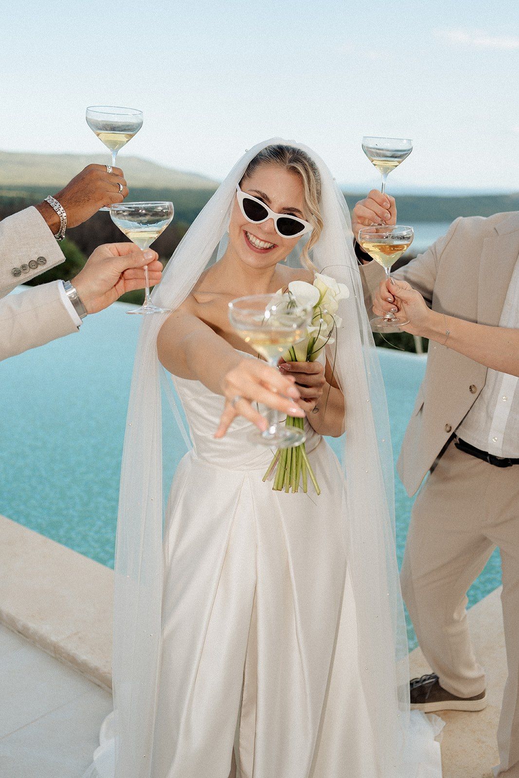 Smiling bride in sunglasses toasts with champagne glasses surrounded by wedding guests outdoors.