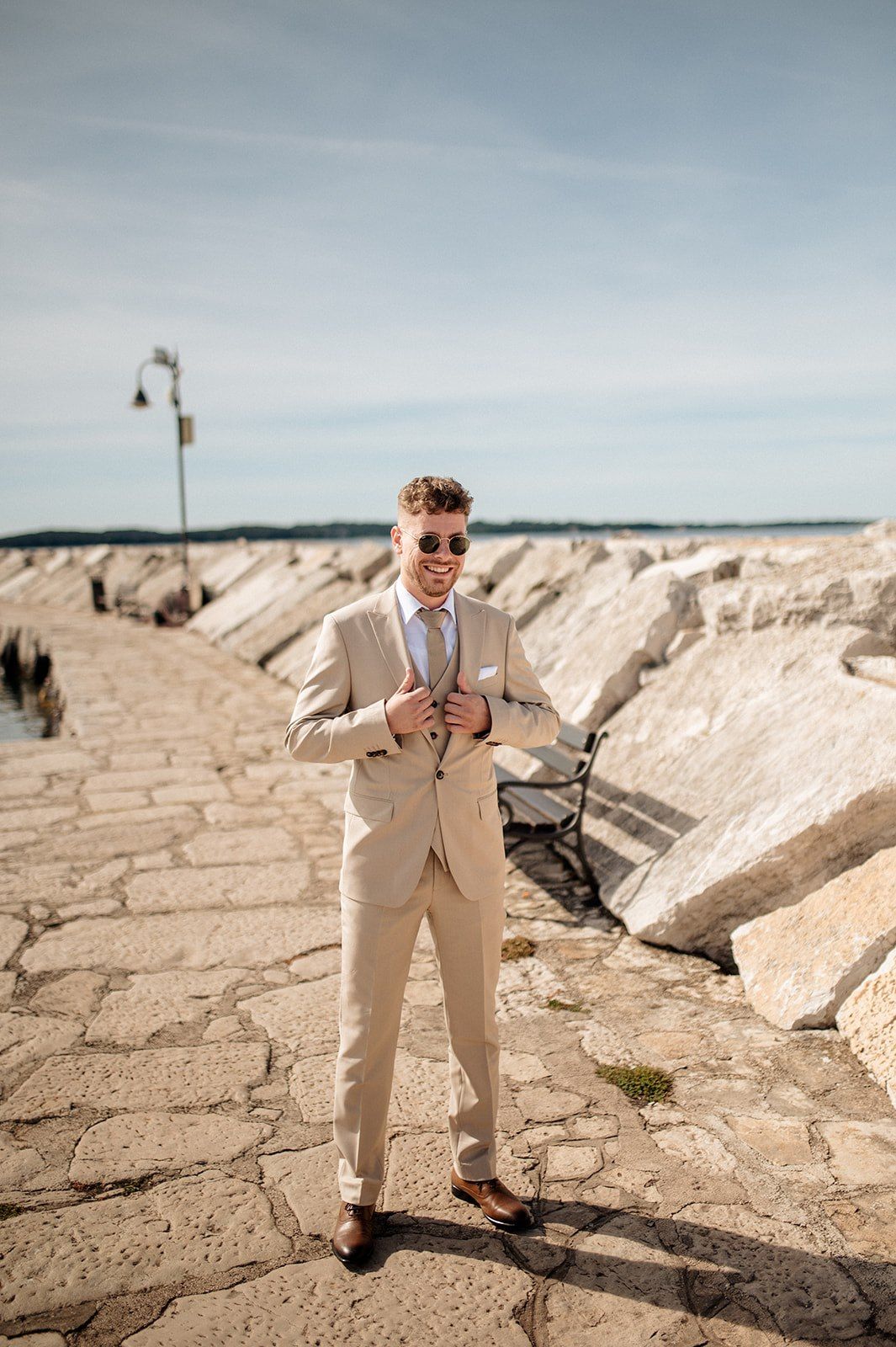 Man in beige suit and sunglasses posing on rocky waterfront pier on a sunny day.