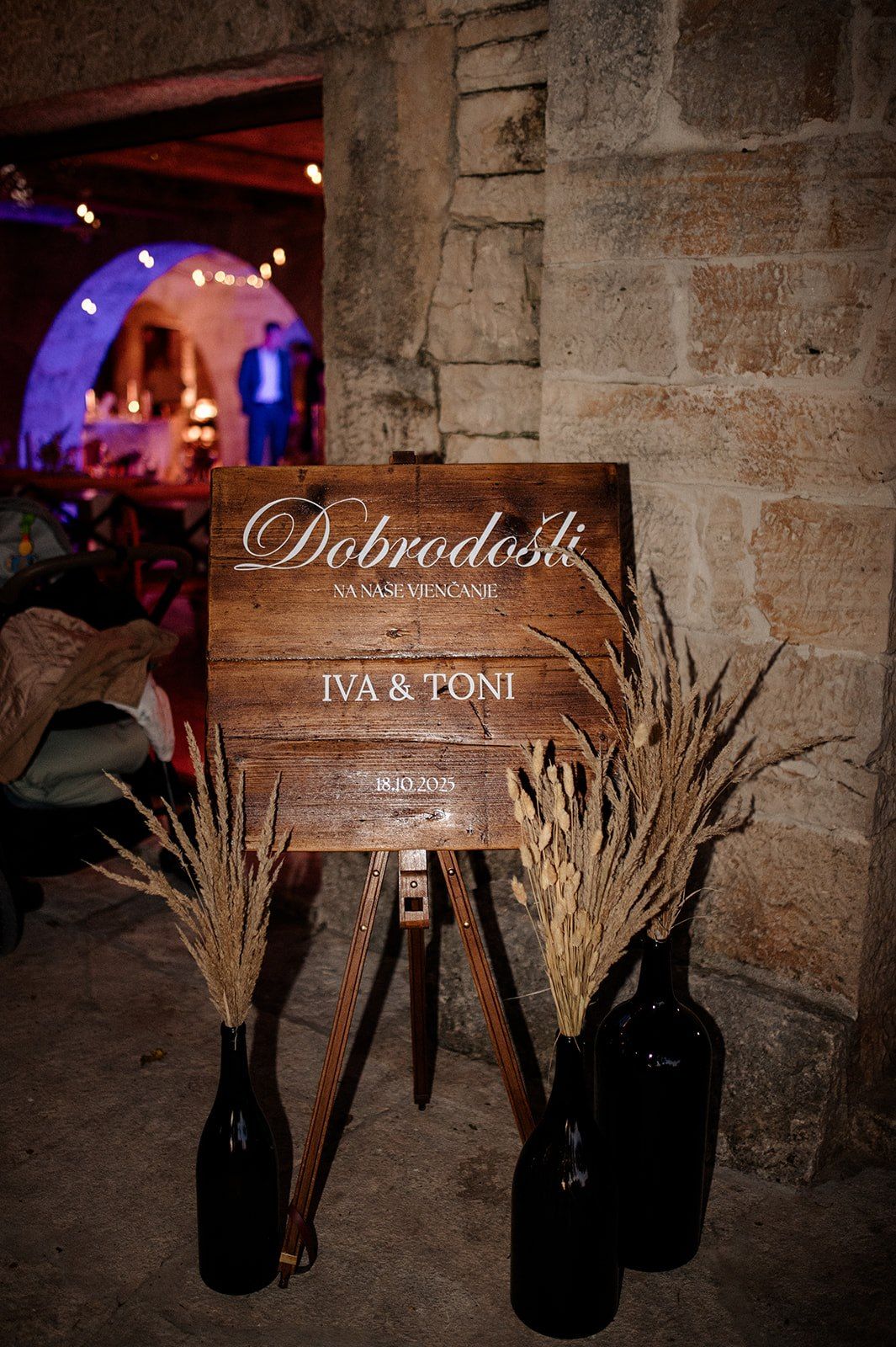 Wooden welcome sign reading Dobrodosli for wedding of Iva and Toni, flanked by dried pampas grass in dark bottles.