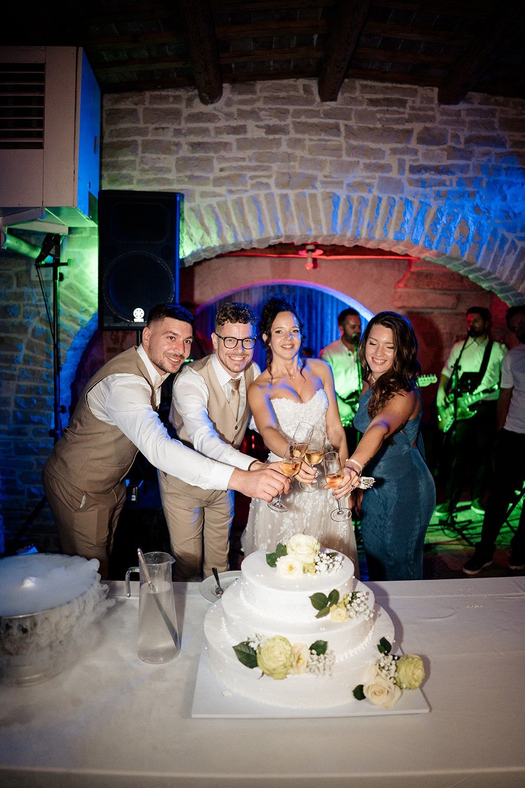 Wedding couple and guests toasting with champagne glasses over a white tiered cake in a stone venue with colorful lights.