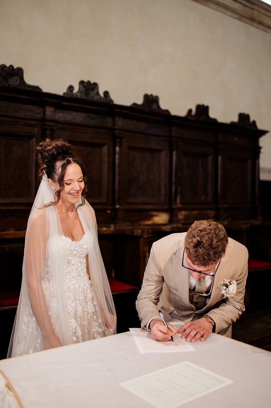 Bride smiling as groom signs wedding certificate at elegant wood-paneled venue during marriage ceremony.