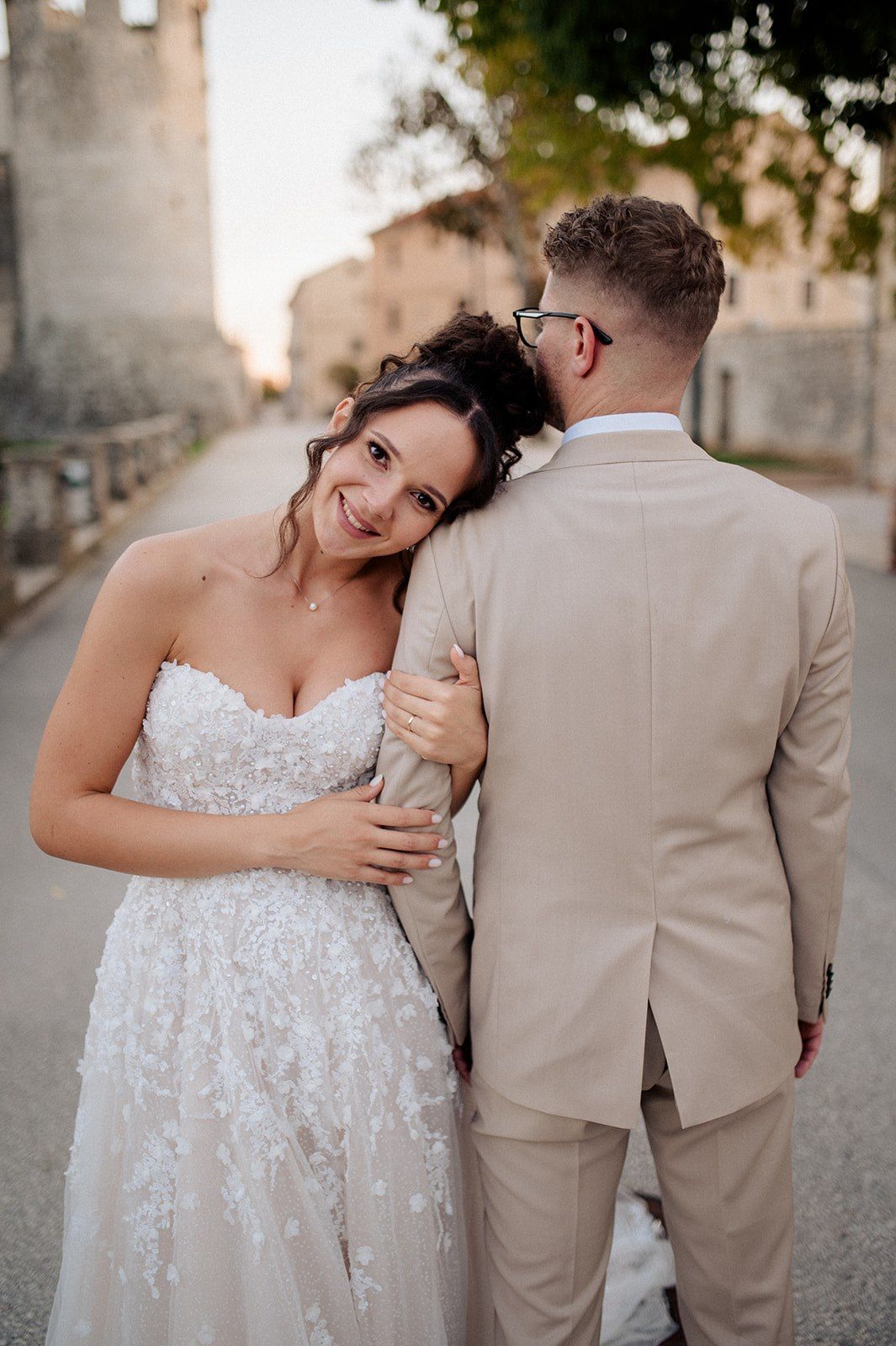 Bride in lace gown leans on groom in beige suit, smiling at camera on a sunlit European street.