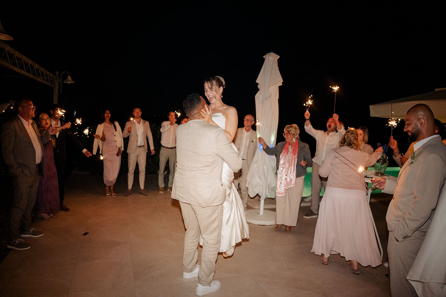 Bride and groom share a joyful first dance surrounded by guests holding sparklers at an outdoor evening wedding reception.