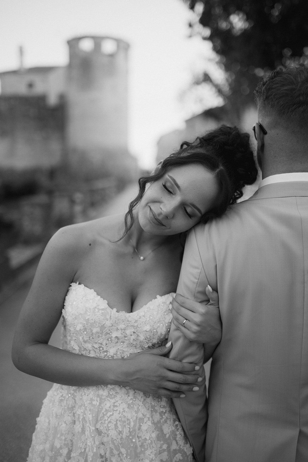 Black and white wedding photo of bride leaning on groom's shoulder, wearing strapless lace gown, with historic tower backdrop.