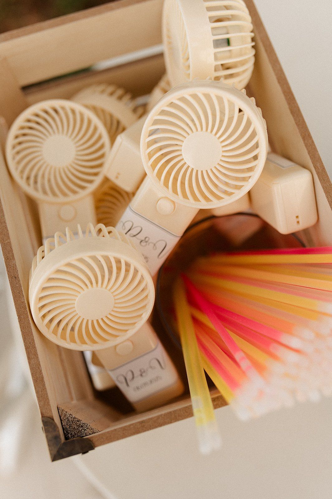 Cream handheld mini fans with monogram labels displayed in a wooden box alongside colorful paper straws as wedding favors.