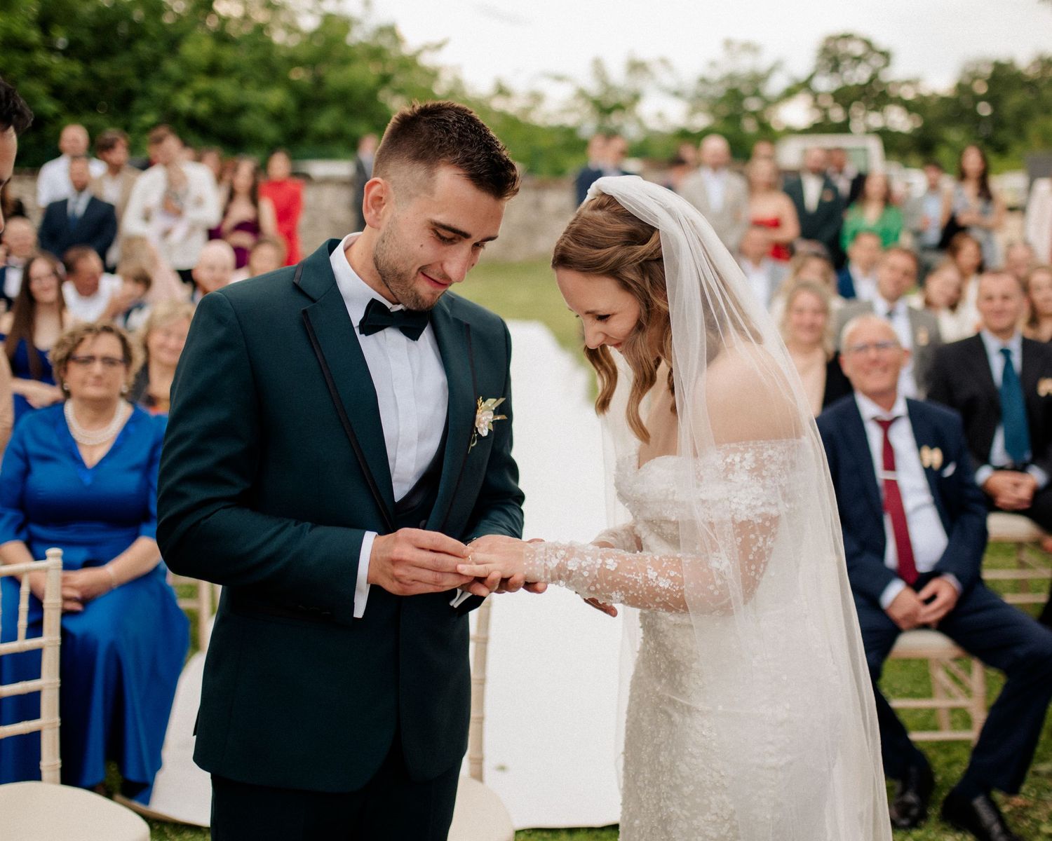 An elegant outdoor wedding ceremony with guests seated on white chairs while a couple exchanges vows beneath floral decor.