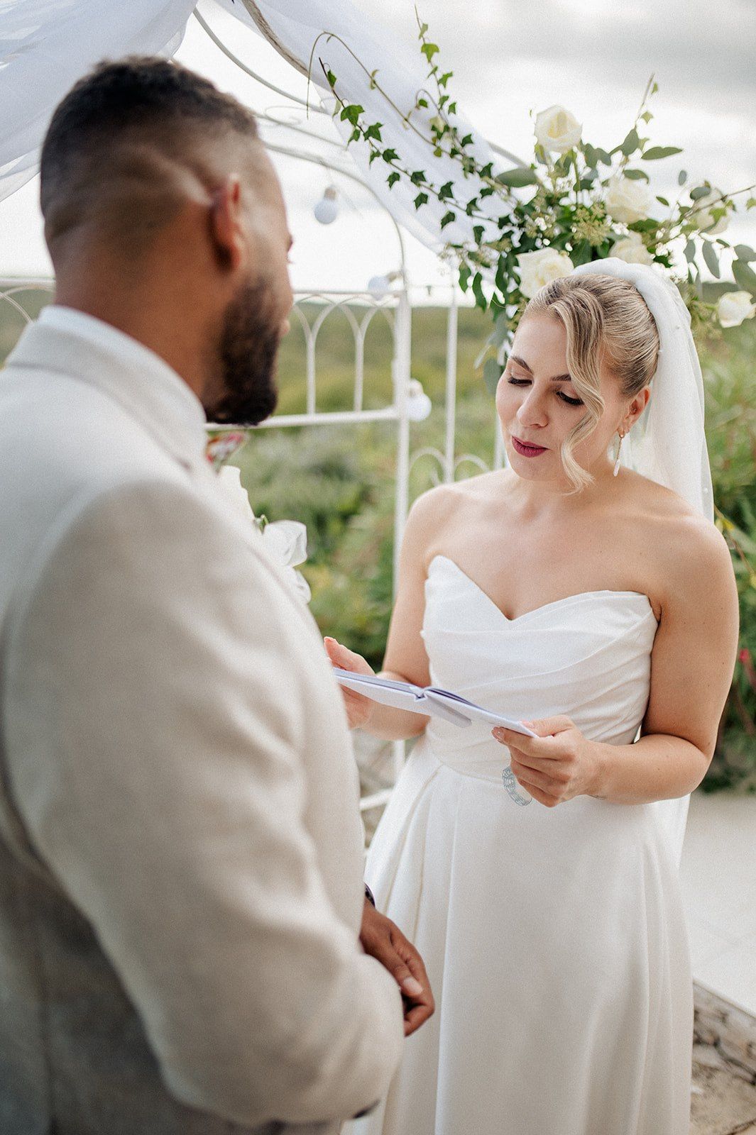 Bride in strapless white gown reads vows to groom during outdoor wedding ceremony beneath floral arch.