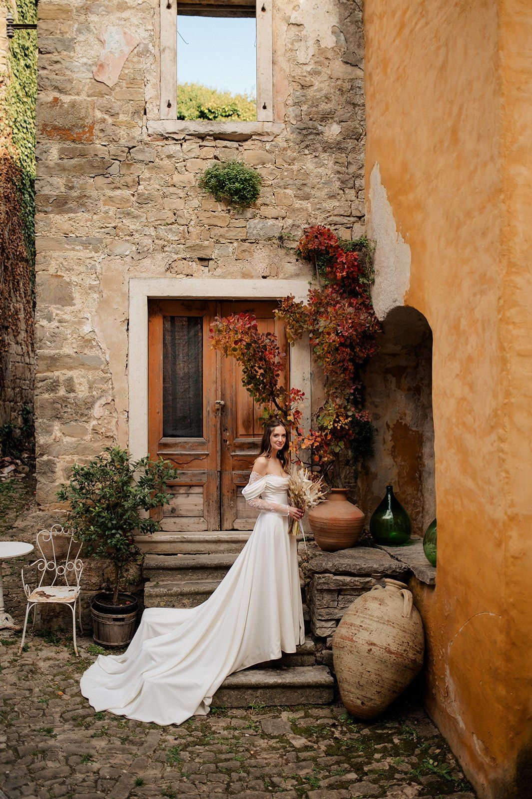 Bride in white off-shoulder gown poses on rustic stone steps of a Tuscan villa with autumn foliage.