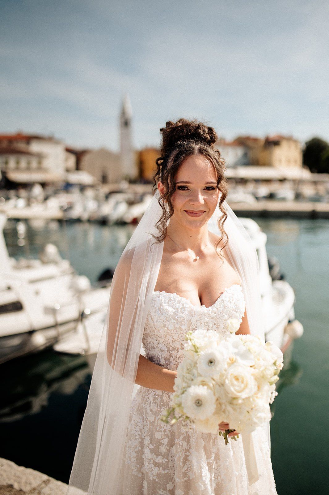 Elegant bride in lace gown holds white floral bouquet at a sunny Croatian marina with boats and a church tower.