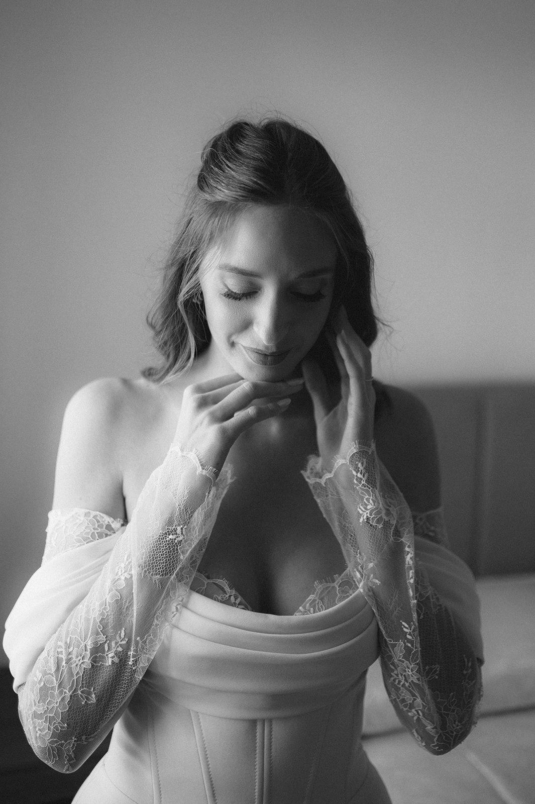 Black and white bridal portrait of a woman in lace sleeves and corset gown, smiling softly with hands near her face.