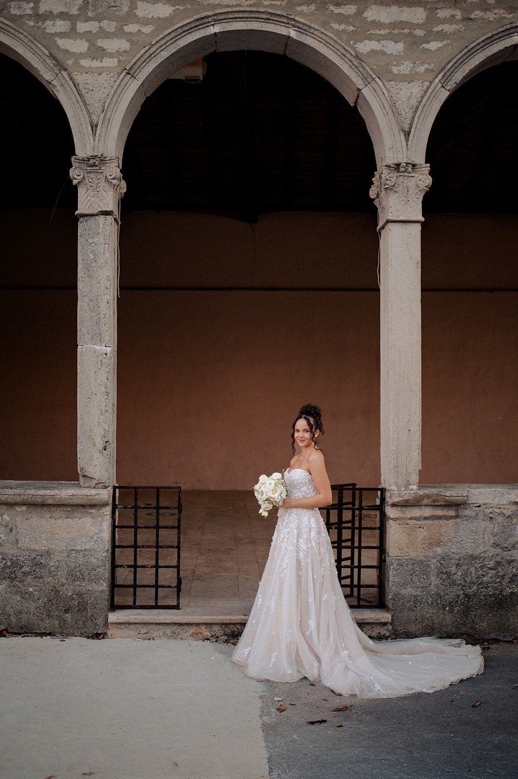 Bride in white lace gown holding bouquet stands under stone arches of elegant historic venue.
