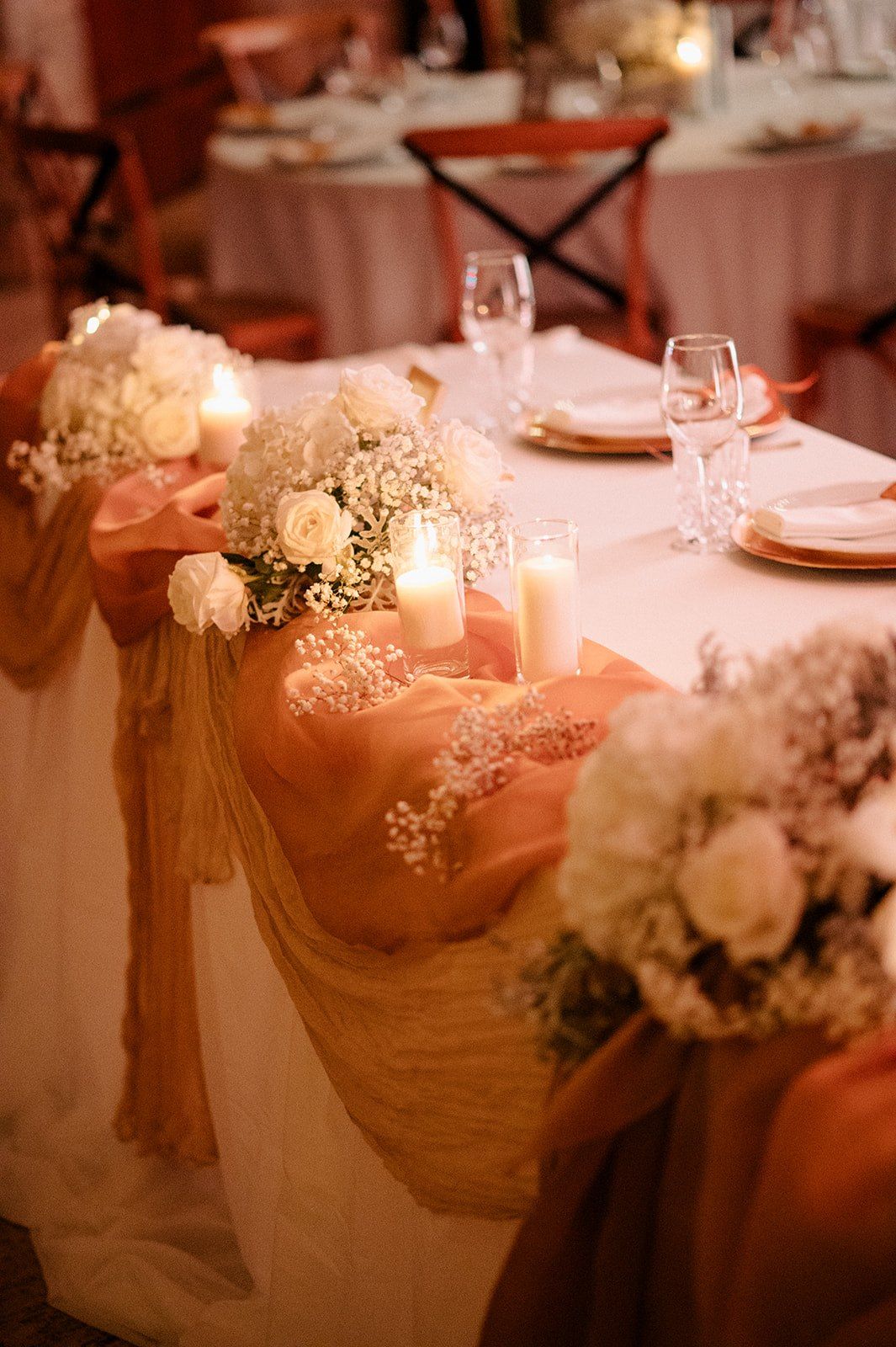 Elegant wedding reception table with white roses, baby's breath, candles, and peach organza fabric draping.