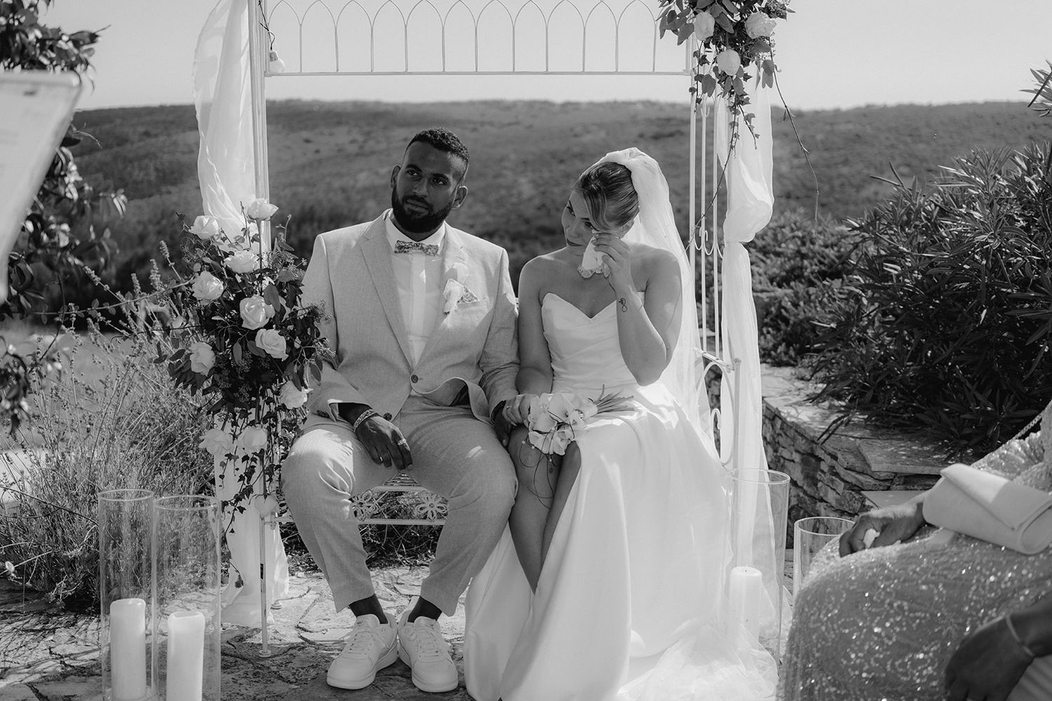 Black and white photo of a bride and groom sitting together at an outdoor wedding ceremony with floral arch.
