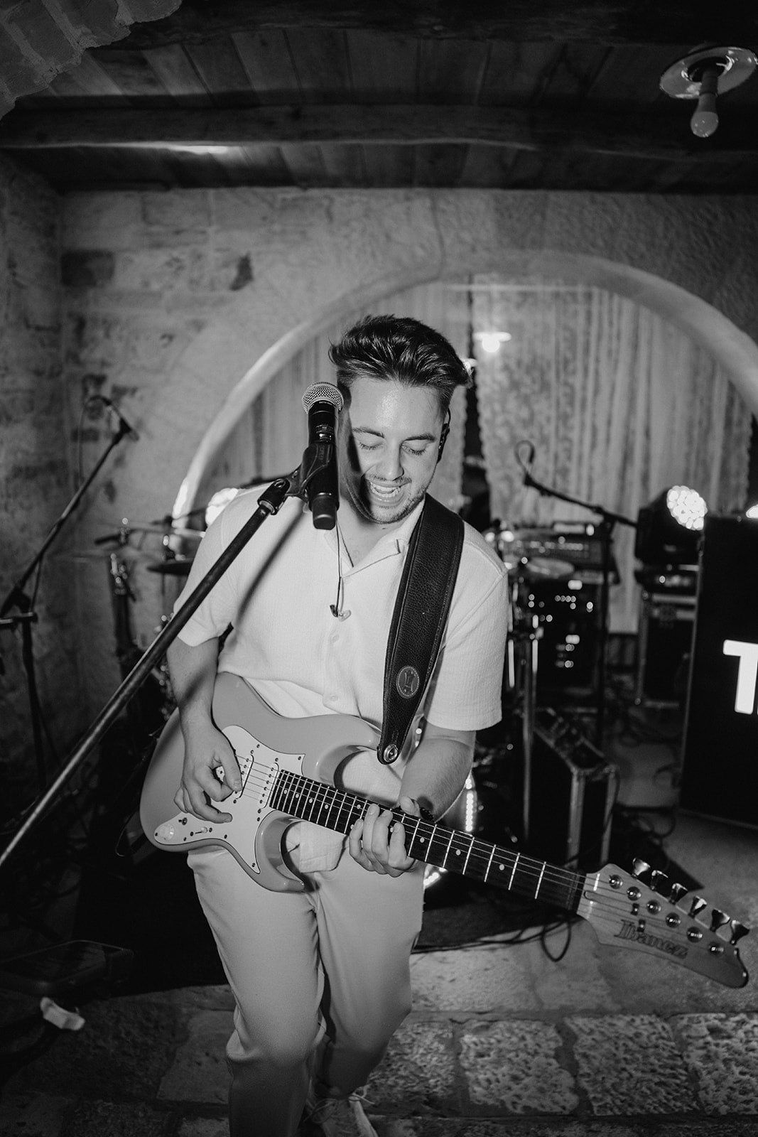 Black and white photo of a young male guitarist smiling while playing electric guitar on stage at a live venue.