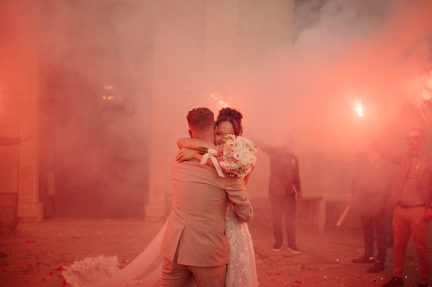 Bride and groom embrace surrounded by red smoke flares during romantic outdoor wedding celebration.
