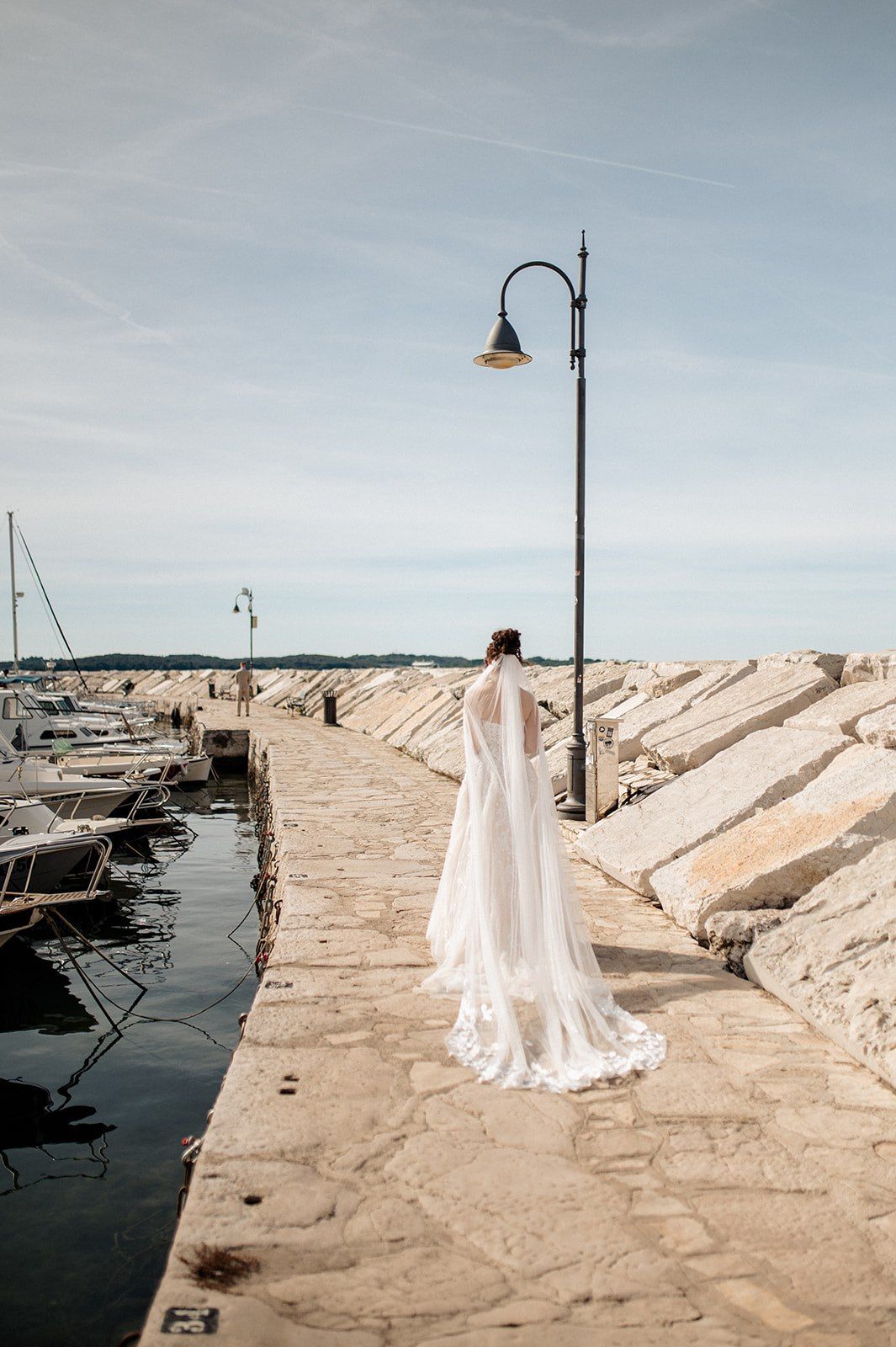 Bride in white wedding gown and veil stands on a stone harbor pier beside a lamp post with boats docked nearby.