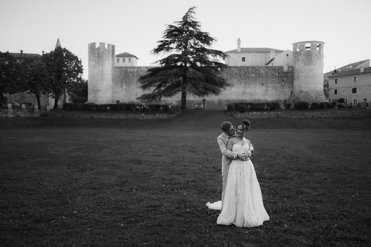 Black and white wedding photo of bride and groom embracing in front of a historic castle surrounded by trees.