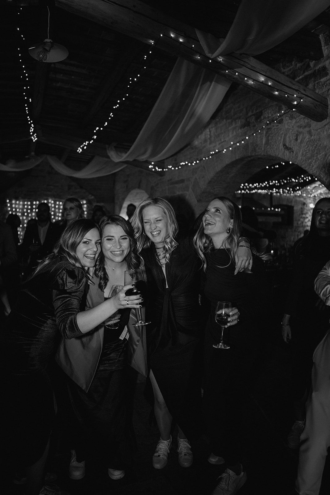 Four women smiling and posing together at an indoor evening event decorated with string lights and draped fabric.