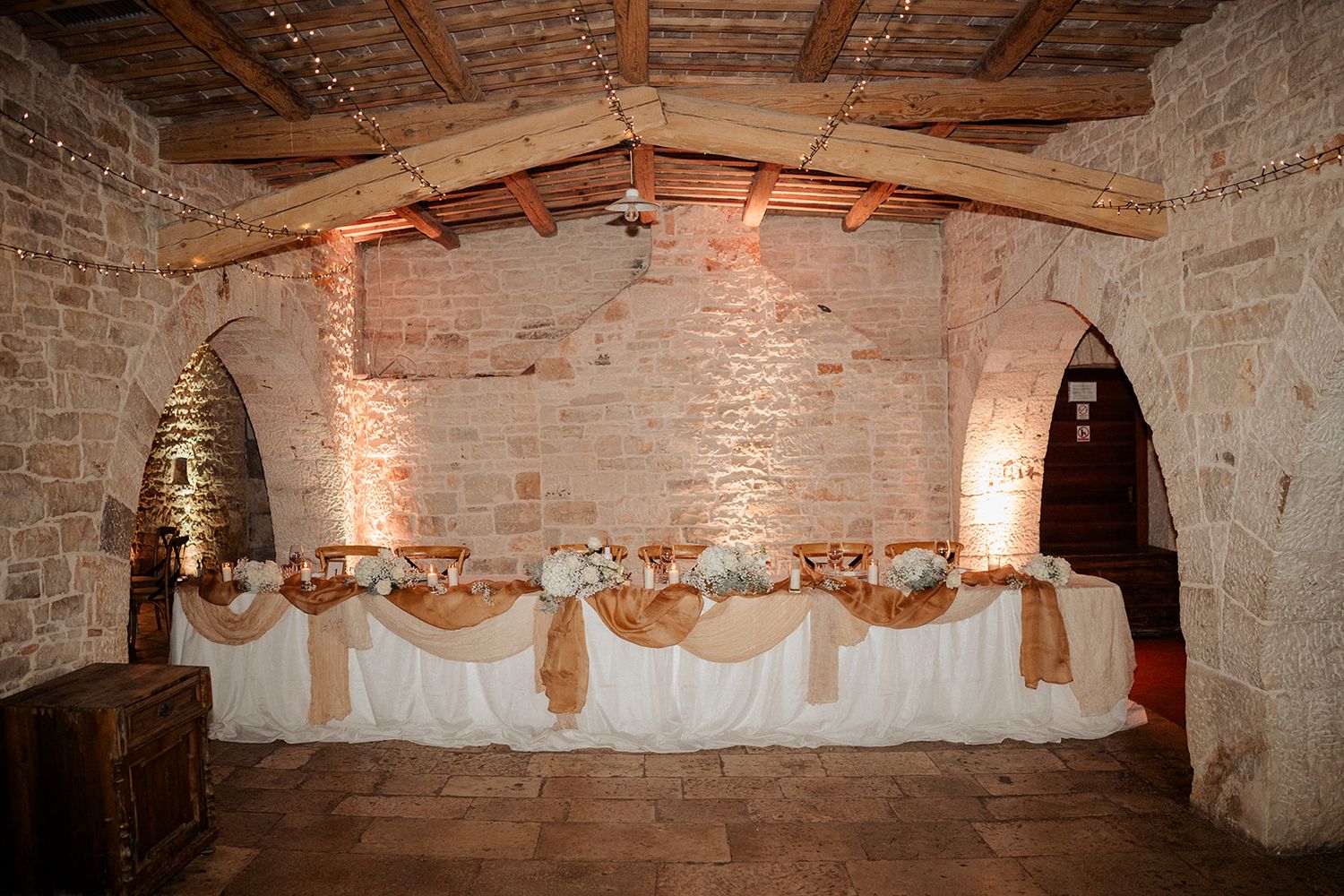 Rustic stone venue with elegant draped head table, wooden beams, and warm amber uplighting for a wedding reception.